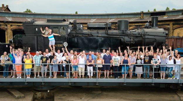 Große Gruppe von Lokführern stehen winkend mit Ihren Partnern und Familie aufgereiht vor einer alten Dampflok